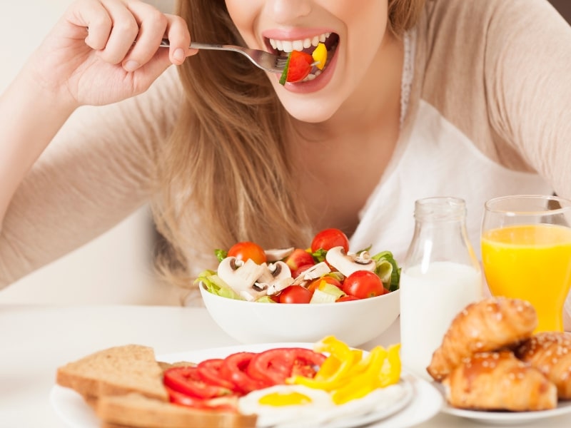 Smiling woman enjoying a healthy breakfast, highlighting her front dental implant while eating fresh vegetables and juice.