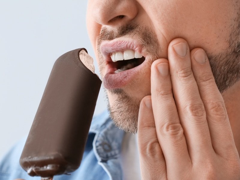 Man experiencing tooth sensitivity pain while eating an ice cream bar, holding his jaw in discomfort.