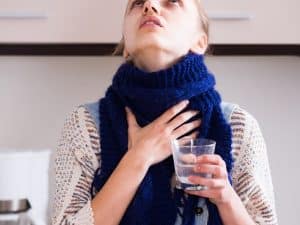 Woman holding her throat and a glass of water, possibly experiencing discomfort related to bleeding gums when brushing.