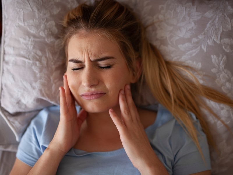 Woman in discomfort holding her jaw while lying down, experiencing sensitivity. Understanding why teeth feel rough overnight.