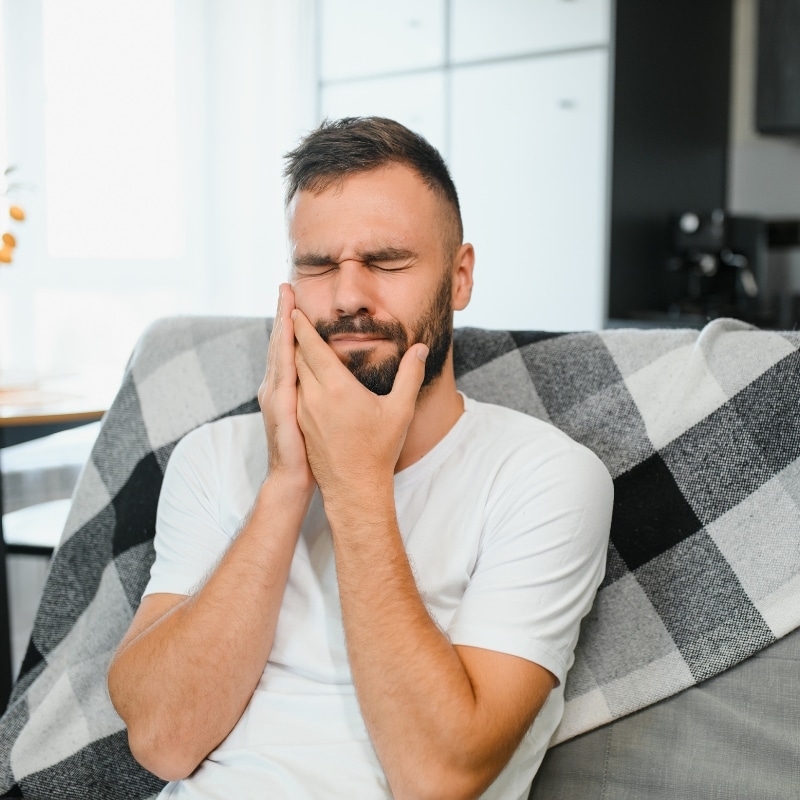 Teeth Whitening sensitivity shown by a man holding his jaw in pain while sitting on a couch at home.